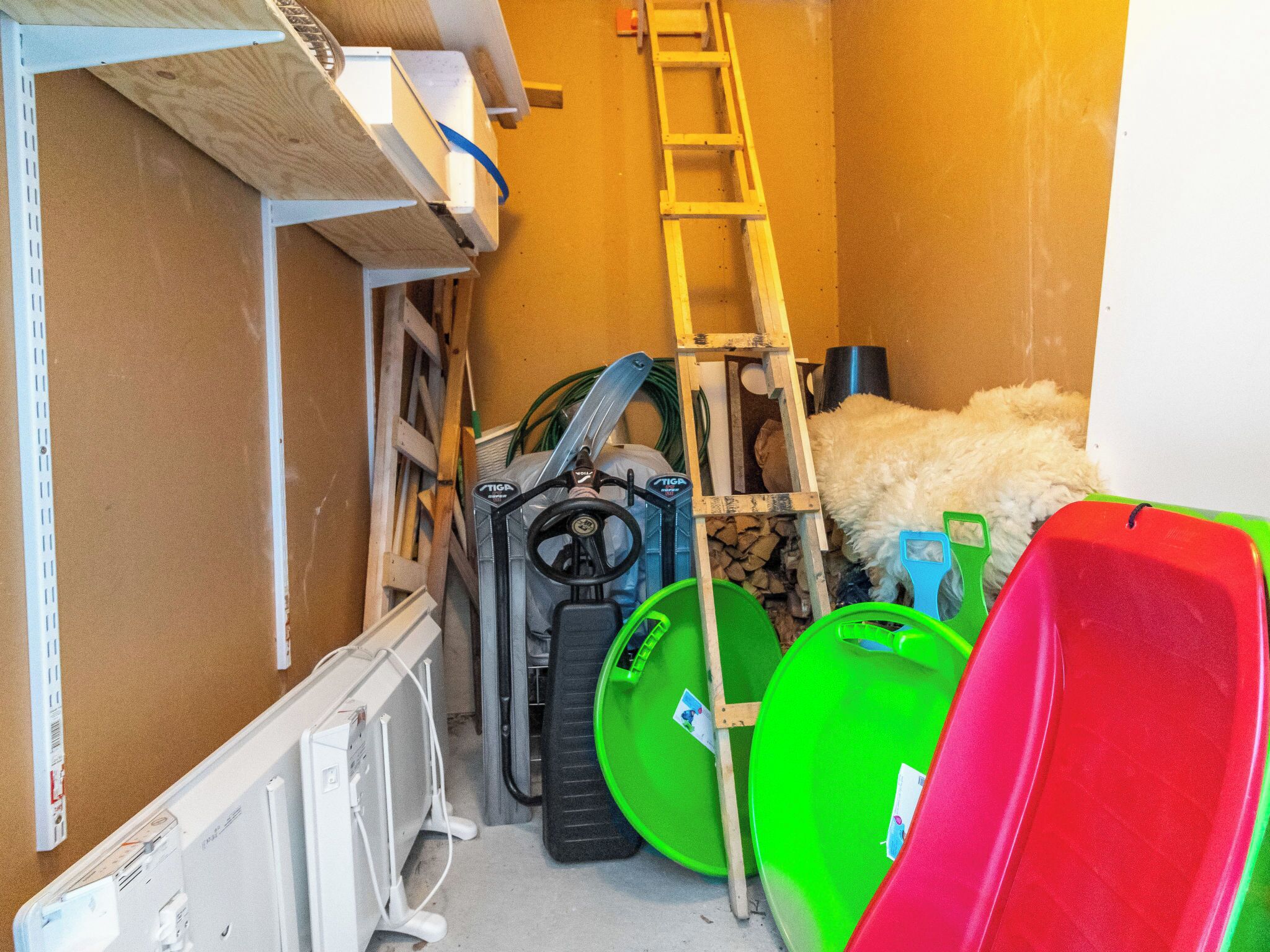 A storage area with various items including a ladder, wheelbarrows, and a pile of wood.