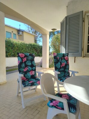 An outdoor patio with two white chairs and a table, surrounded by a beige wall and a window with shutters. - Cottage with large patio and bike path that connects directly to the waterfront (Cecina)