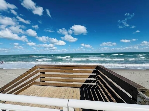 Une terrasse en bois avec une rambarde, donnant sur une plage où l’on peut admirer les vagues et un ciel bleu dégagé.