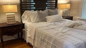 Een slaapkamer met een houten hoofdbord, twee nachtkastjes met lampen, een bed met een decoratief sprei en een raam met luxaflex.