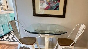 Een eethoek met een glazen tafel en twee witte stoelen, een wand waarop een abstract schilderij hangt, en een keuken met teal kasten.