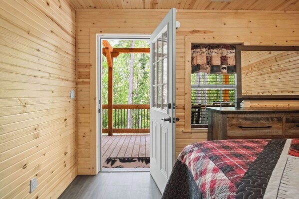 A wooden cabin interior with a bed, a wooden dresser, a door leading to a balcony, and a window with curtains.
