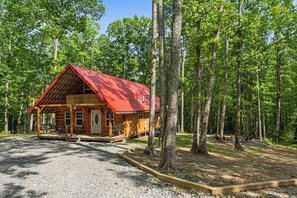 A cabin with a red roof, surrounded by trees, and a gravel driveway.
