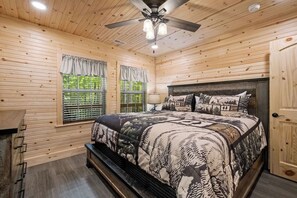 A wooden cabin bedroom with a bed, a ceiling fan, and a window with curtains.