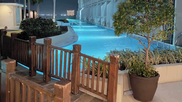 A nighttime view of a hotel pool area with a wooden fence, potted plants, and a multi-story building with balconies. - SkyLine CtyView Condotel BREEZE RESIDENCES near MOA,NAIA AIRPORT,PICC,US Embassy (Pasay City)