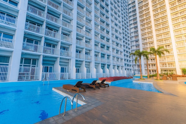 A large hotel with multiple balconies and a swimming pool area with lounge chairs.
