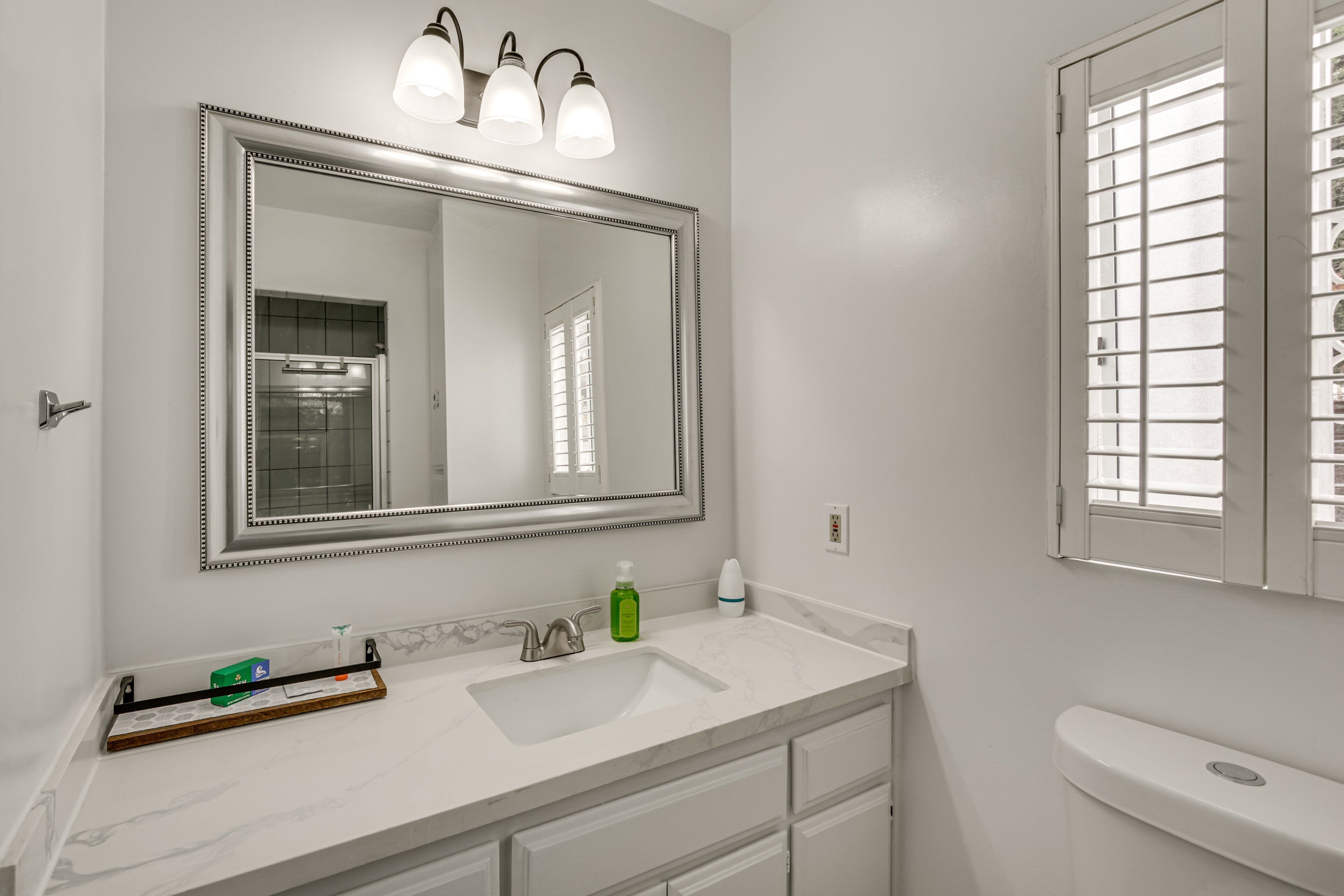 A bathroom with a white vanity, a large mirror with three lights, a toilet, and a window with blinds.