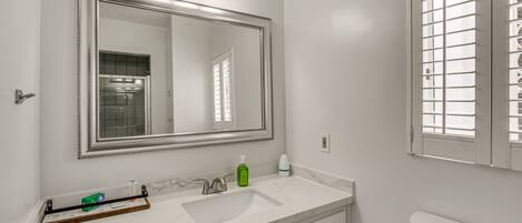 A bathroom with a white vanity, a large mirror with three lights, a toilet, and a window with blinds.