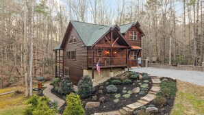 A wooden cabin with a balcony, surrounded by a landscaped garden and a forested area. - Spacious Smoky Mountain cabin with fire pit, hot tub, and game room (Sevierville)