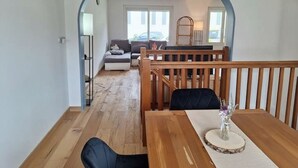 Un comedor con una mesa y sillas de madera, un florero con flores, y vista a una sala de estar con un sofá y un piano.
