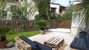 Un patio con mobiliario de madera, incluyendo un sofá y un banco, rodeado por un cerco de madera y macetas con plantas.