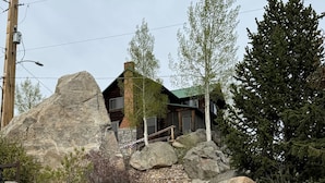 Eine Holztreppe führt zu einer Hütte mit einer Steinwand, umgeben von großen Steinen und Bäumen.