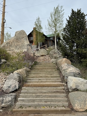 Un escalier en bois mène à un chalet dont un des murs est en pierre, et qui est entouré de gros rochers et d’arbres.