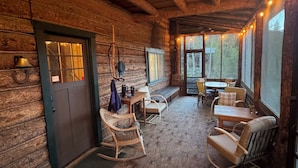 Ein rustikales Blockhaus-Interieur mit Holzwänden, einer Essecke mit Stühlen und Tisch sowie einem großen Fenster mit Blick ins Freie.