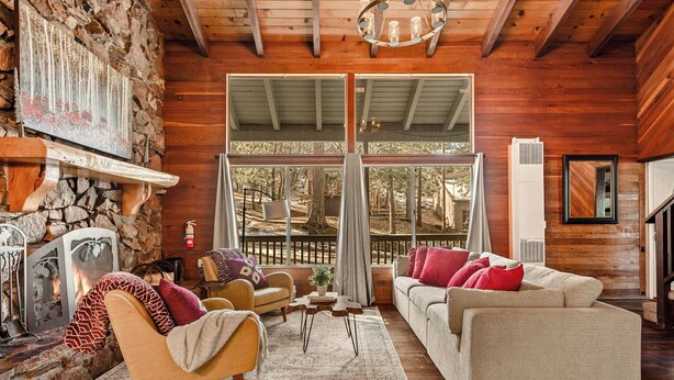 A cozy living room with a stone fireplace, wooden beams, and a large window overlooking a deck. - Stylish & Cozy "Nut House" - Great & Game Room - Spa (Big Bear)
