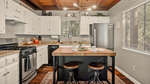 A modern kitchen with white cabinets, a central island with stools, a stainless steel refrigerator, and a wooden ceiling. - Stylish & Cozy "Nut House" - Great & Game Room - Spa (Big Bear)