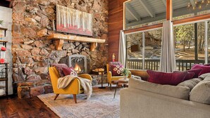 A cozy living room with a stone fireplace, wooden beams, and large windows overlooking a deck. - Stylish & Cozy "Nut House" - Great & Game Room - Spa (Big Bear)