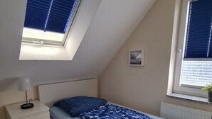 A bedroom with a bed, a nightstand, a lamp, a window with blinds, a radiator, and a striped rug. - Cozy apartment Strandallee in Burg/Fehmarn. (Burg/Fehmarn)