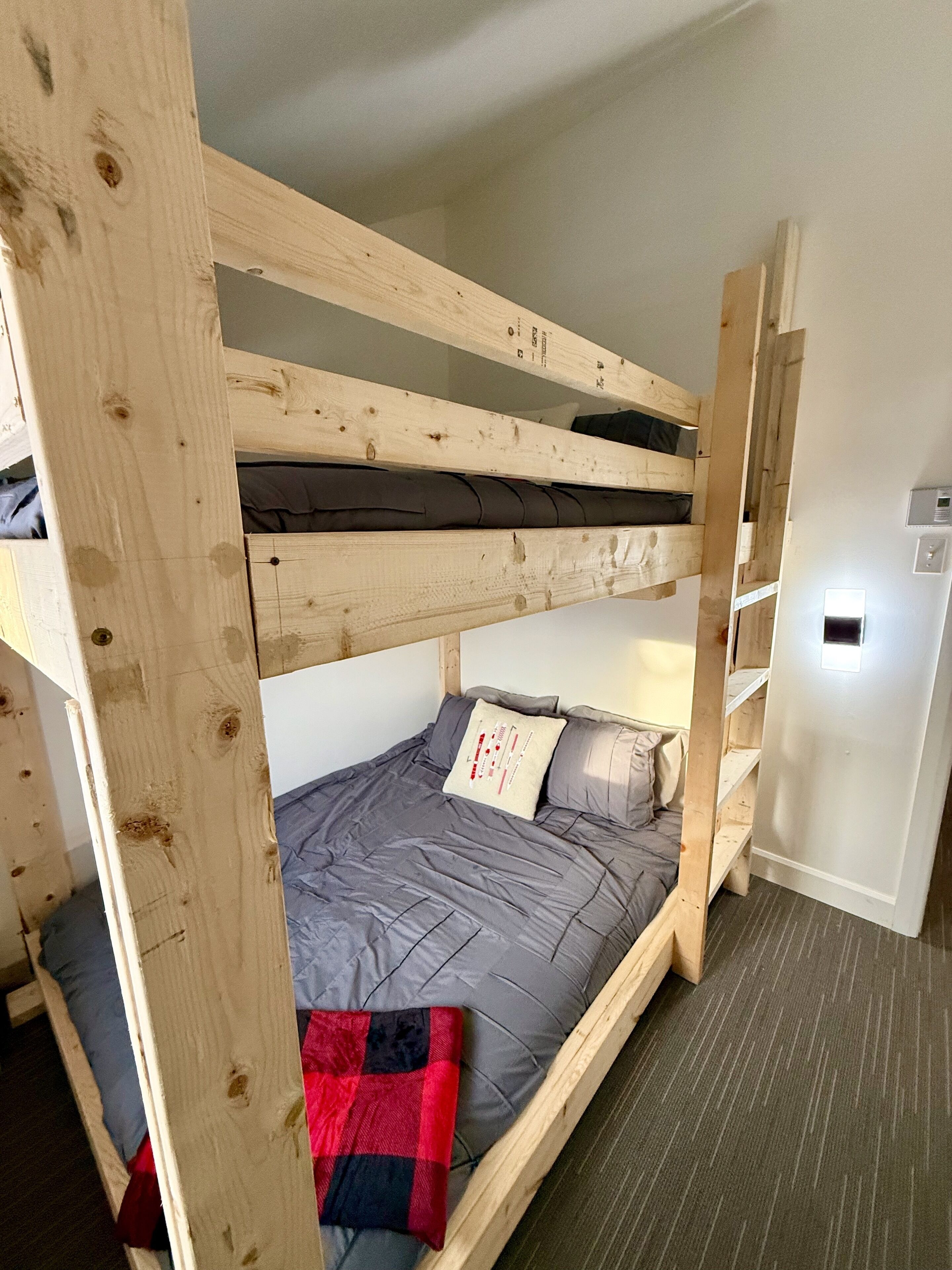 A bunk bed with a grey mattress and a red and black blanket, wooden headboard, and a white wall with a thermostat.