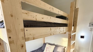 Ein Etagenbett mit grauem Matratze und einer Decke in Rot und Schwarz, ein Holz-Kopfteil und eine weiße Wand mit Thermostat.