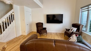 Ein geräumiges Wohnzimmer mit großem Flachbildfernseher an der Wand, einem braunen Ledersofa, zwei Sesseln und einer Treppe mit weißen Geländern.
