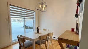 Ein Essbereich mit einem weißen Tisch und Stühlen, einer Hängelampe und Blick durch ein Fenster mit Jalousien.