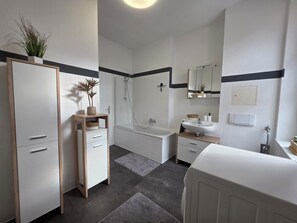 A modern bathroom with a bathtub, shower, and sink. There is a wooden cabinet with drawers and a potted plant. - Cozy 3-room apartment near the center (Magdeburg)