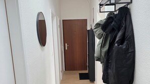 Un pasillo con suelo de madera, un banco negro, un perchero con abrigos, una planta en maceta y una puerta al final.