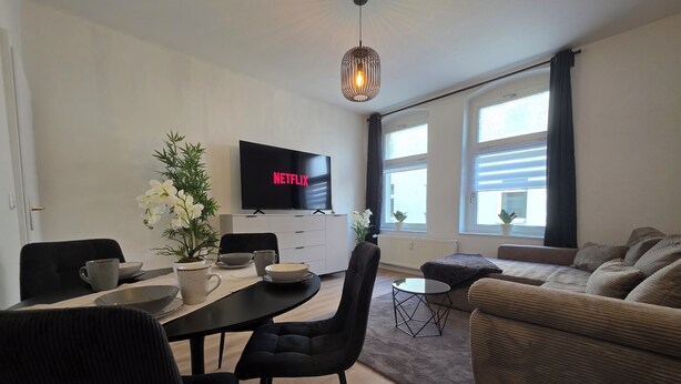 Una sala de estar moderna con comedor, un televisor de pantalla plana mostrando 'NETFLIX', un sofá y una mesa de centro.