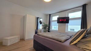 Un dormitorio con cama, televisión montada en la pared, ventana y planta.