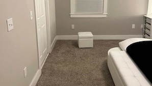 Un dormitorio con una cama individual, una ventana con persianas, una puerta y un pequeño reposapiés blanco.