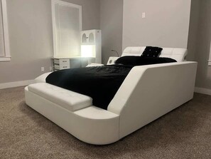 A bedroom with a bed, a bedside table, a lamp, a window with blinds, and a wall-mounted heater.