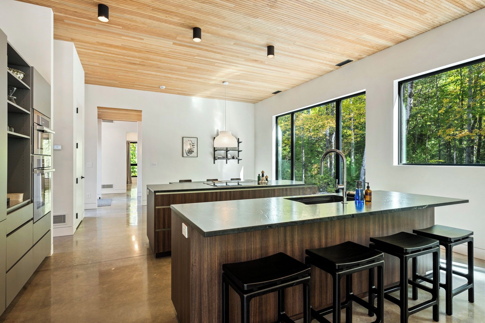 A modern kitchen with a central island, bar stools, and a large window overlooking a forest.
