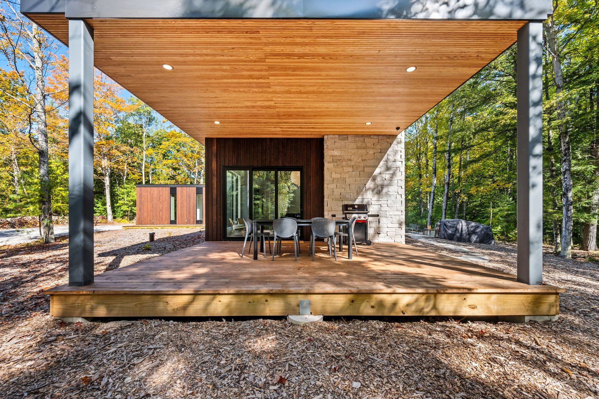 A modern outdoor dining area with a wooden deck, metal chairs, and a stone wall.