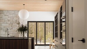 A modern kitchen with a wooden ceiling, stone wall backsplash, and large windows. - Cedar & Glass | Modern Architecture Forest Retreat (Fish Creek)