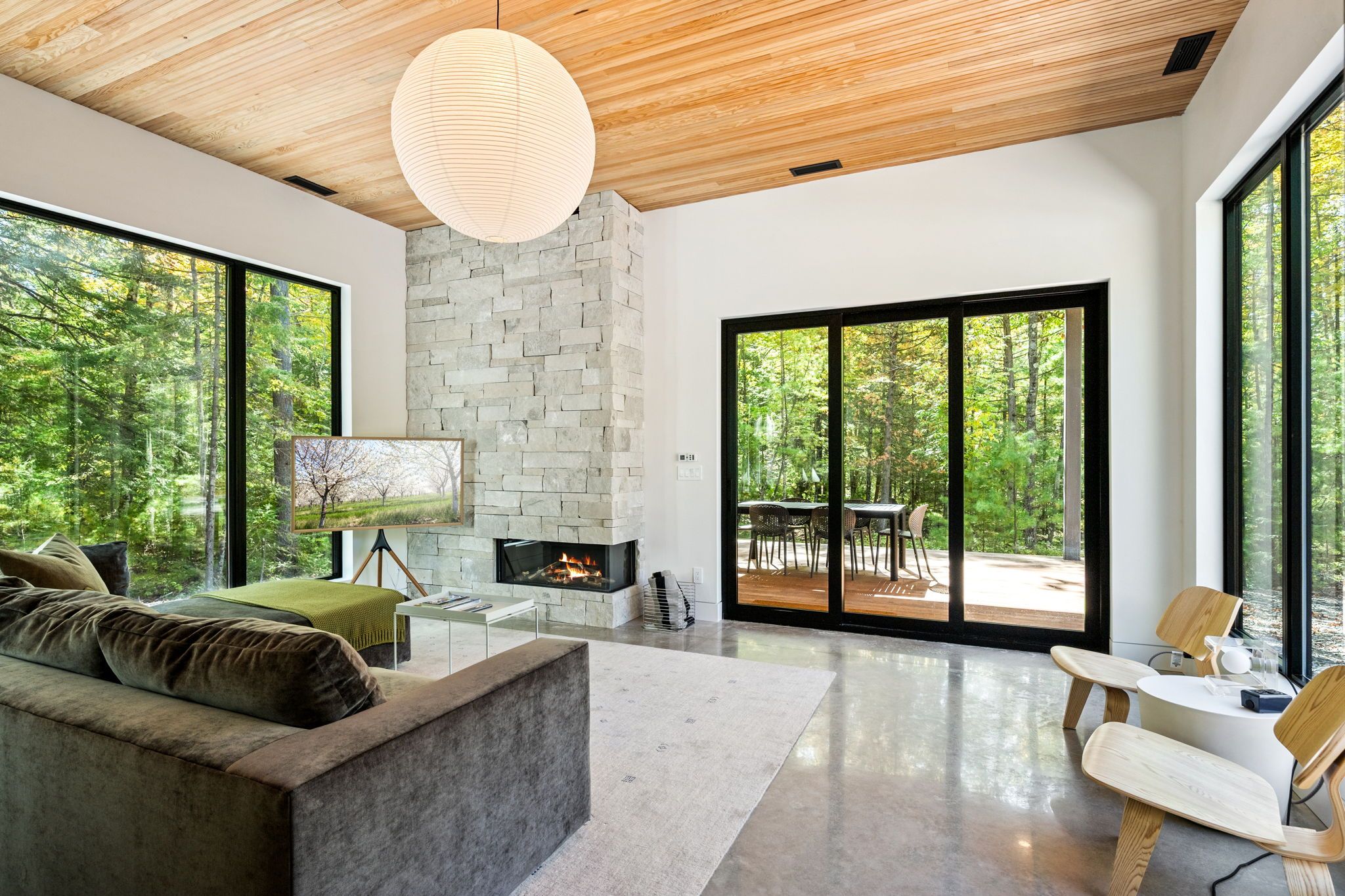 A modern living room with a stone fireplace, large windows, and a view of the forest.