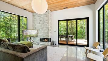 A modern living room with a stone fireplace, large windows, and a view of the forest.
