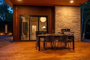 An outdoor dining area with a wooden deck, a stone wall, and a grill. - Cedar & Glass | Modern Fish Creek Forest Retreat (Fish Creek)