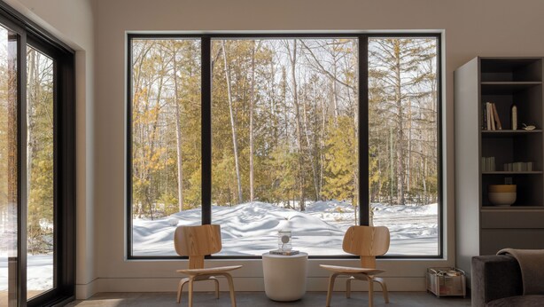 Een moderne woonkamer met een groot raam, twee houten stoelen, een klein tafeltje en een boekenkast.
