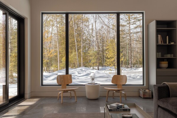 Una sala de estar moderna con un ventanal grande, dos sillas de madera, una mesita y una estantería.
