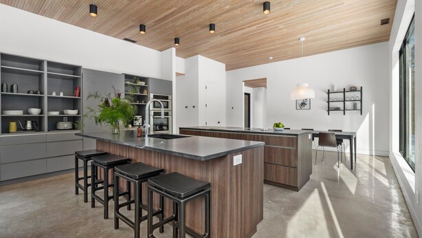 A modern kitchen with a central island, bar stools, and built-in shelves. - Cedar & Glass | Modern Architecture Forest Retreat (Fish Creek)