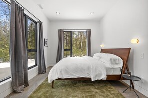 A modern bedroom with a large window, a bed, a small table, and a green rug. - Cedar & Glass | Modern Fish Creek Forest Retreat (Fish Creek)