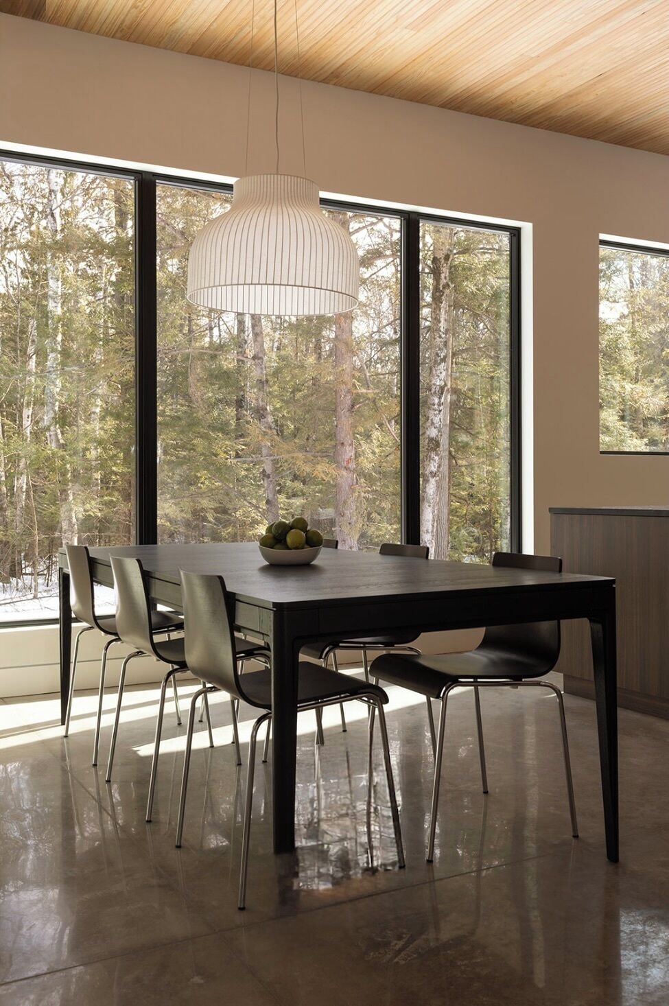A modern dining area with a wooden ceiling, large windows with a forest view, a dark dining table with chairs, and a hanging light fixture.
