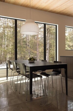 A modern dining area with a wooden ceiling, large windows with a forest view, a dark dining table with chairs, and a hanging light fixture. - Cedar & Glass | Modern Fish Creek Forest Retreat (Fish Creek)
