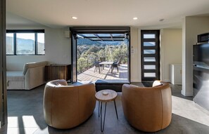 A modern living room with a sofa, two armchairs, a small round table, and a large window overlooking a deck with a scenic view. - PINE unCONTAINED - Modern container home with a wildlife pond & mountain views! (Pine)