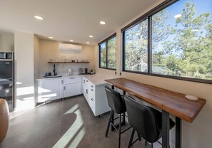 A modern kitchen with a wooden bar, white cabinets, and a large window with a view of trees. - PINE unCONTAINED - Modern container home with a wildlife pond & mountain views! (Pine)