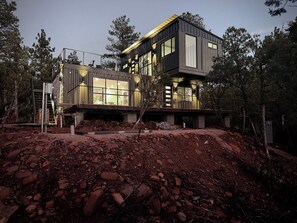 A modern two-story house with large windows, a balcony, and a construction site in the foreground. - PINE unCONTAINED - Modern container home with a wildlife pond & mountain views! (Pine)