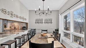 Une cuisine moderne dotée d’un grand îlot central, d’un coin repas avec une longue table et des chaises, et offrant une vue dégagée grâce à de grandes fenêtres.