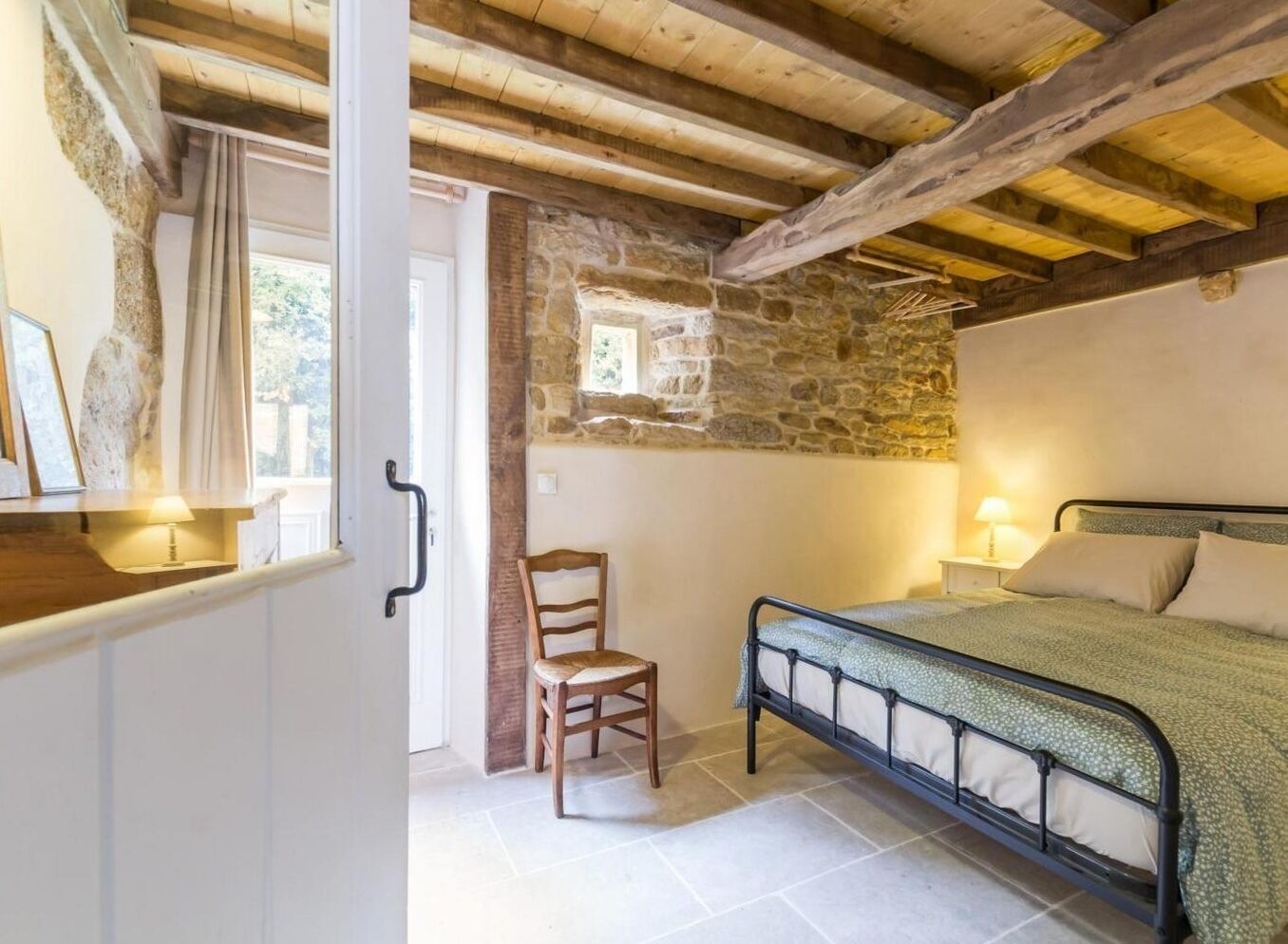 A bedroom with a stone wall, a wooden chair, a bed with a metal frame, and a window with curtains.
