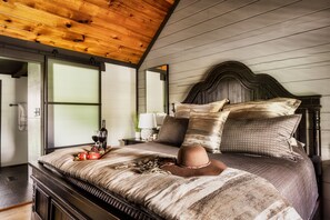 Une chambre à coucher avec un plafond en bois, un lit avec une tête de lit, des oreillers, un chapeau, un plateau avec de la nourriture et du vin, et une table de chevet avec une lampe.